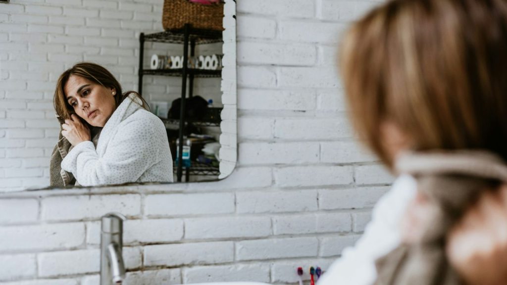 A sad woman in a robe looks in a mirror while drying her hair with a towel.