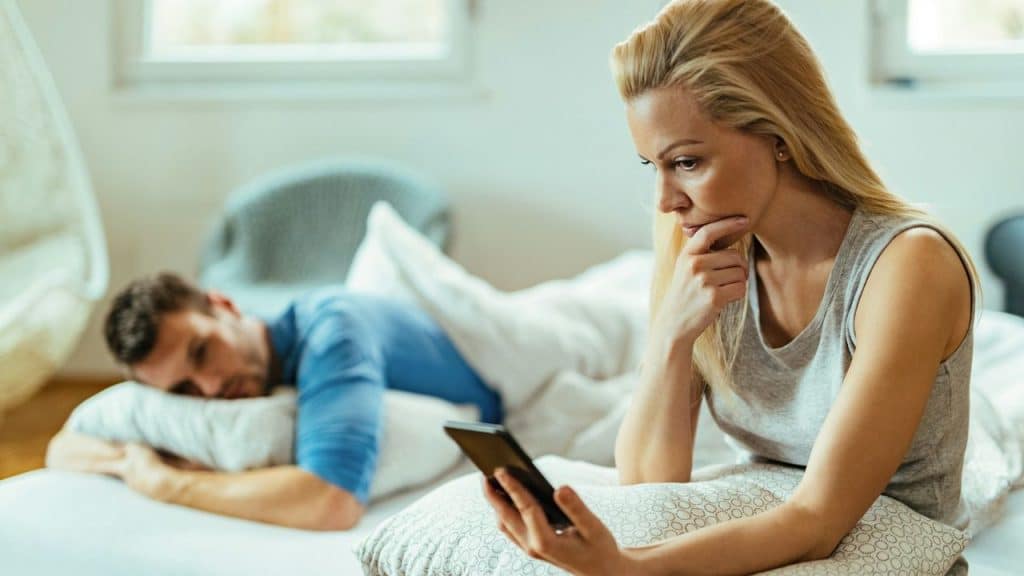 A distraught woman looks at a smartphone while a man sleeps in the background.