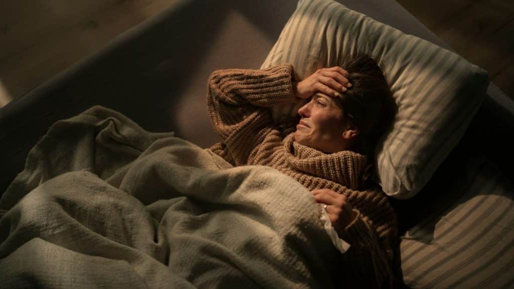 A distraught woman in a cozy sweater lies in bed holding her forehead.