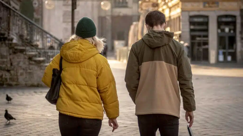A man and woman in jackets walk away from the camera on a city sidewalk in the evening.