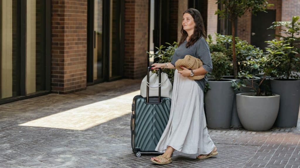 A woman in a long skirt walks with a suitcase and a bag in a courtyard.