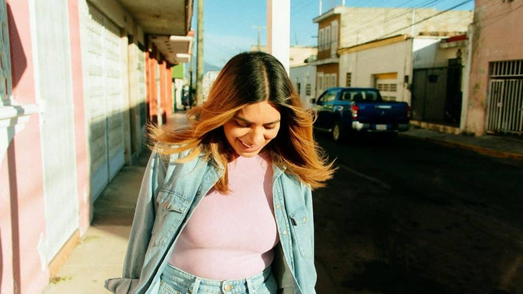 A woman walking on a sunlit street in a denim jacket over a pink top, smiling.