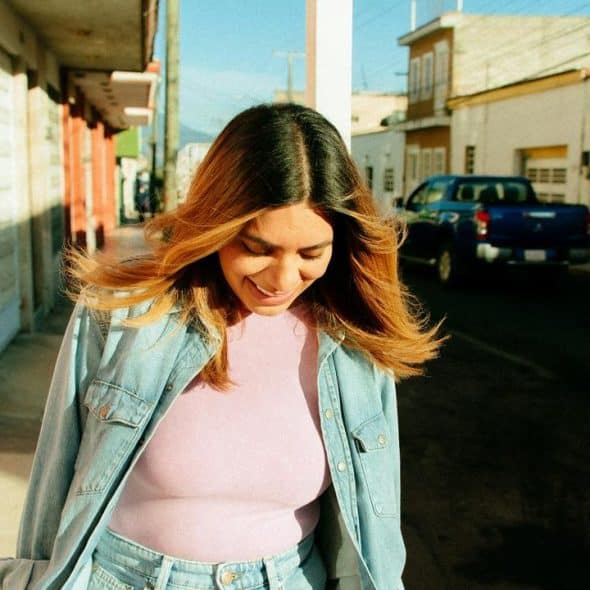 A woman walking on a sunlit street in a denim jacket over a pink top, smiling.
