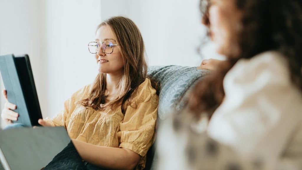 A woman sitting on a couch holding a tablet with another person beside her.
