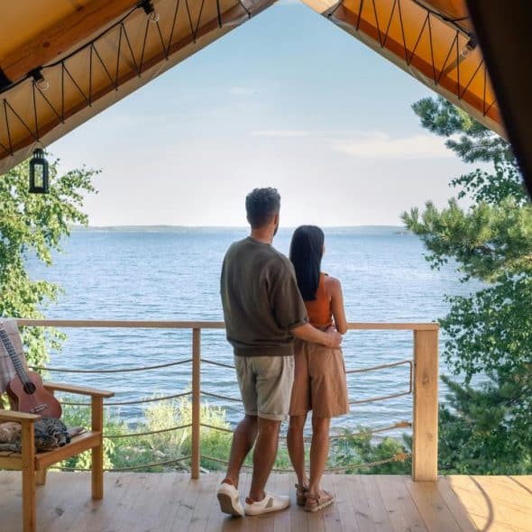 A couple standing on a wooden cabin balcony overlooking a lake.