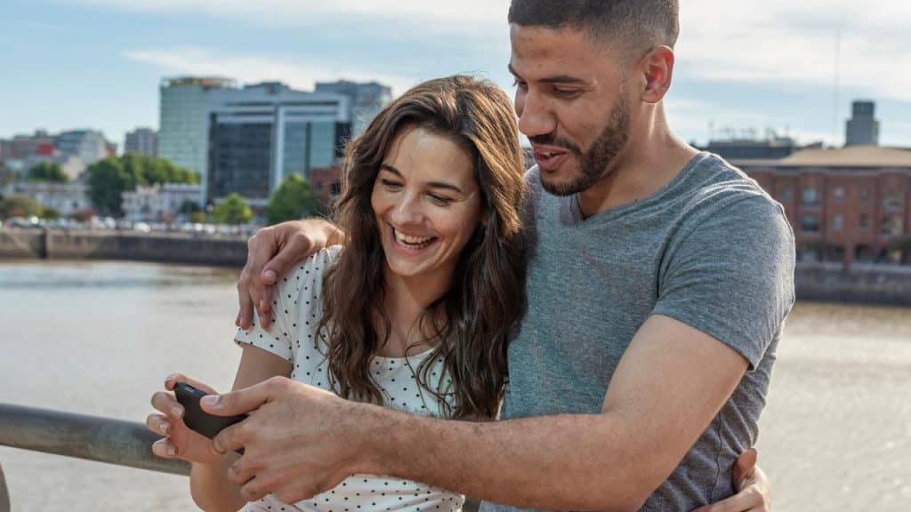 A couple standing by the water smiling at a phone.