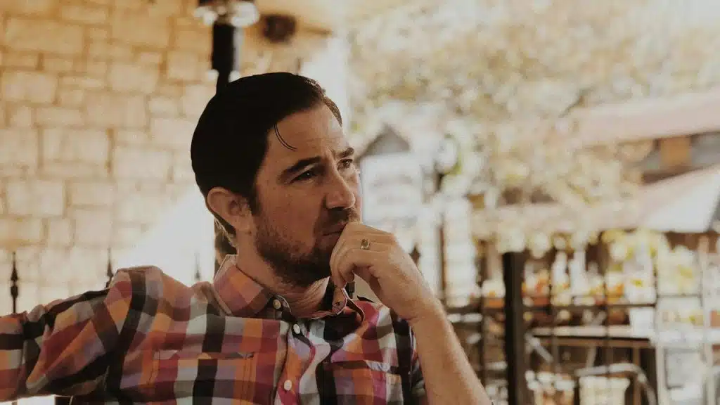 A man in a checkered shirt sitting and looking thoughtful.