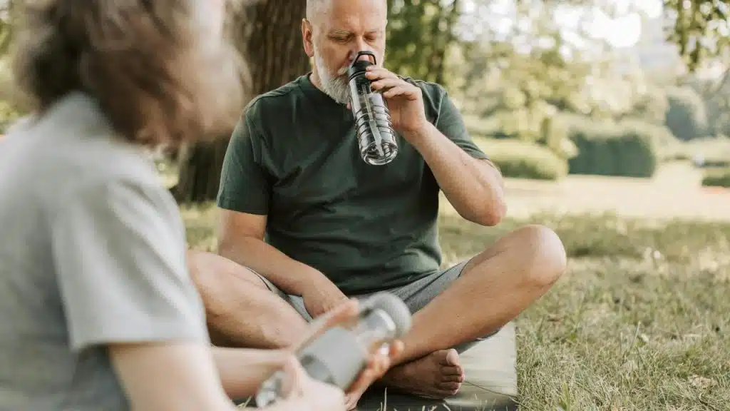 A senior man sitting on the grass drinking water beside a companion.