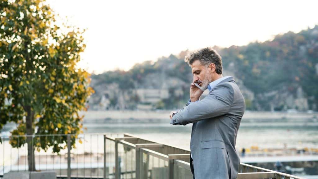 A businessman talking on the phone while checking his watch outdoors.