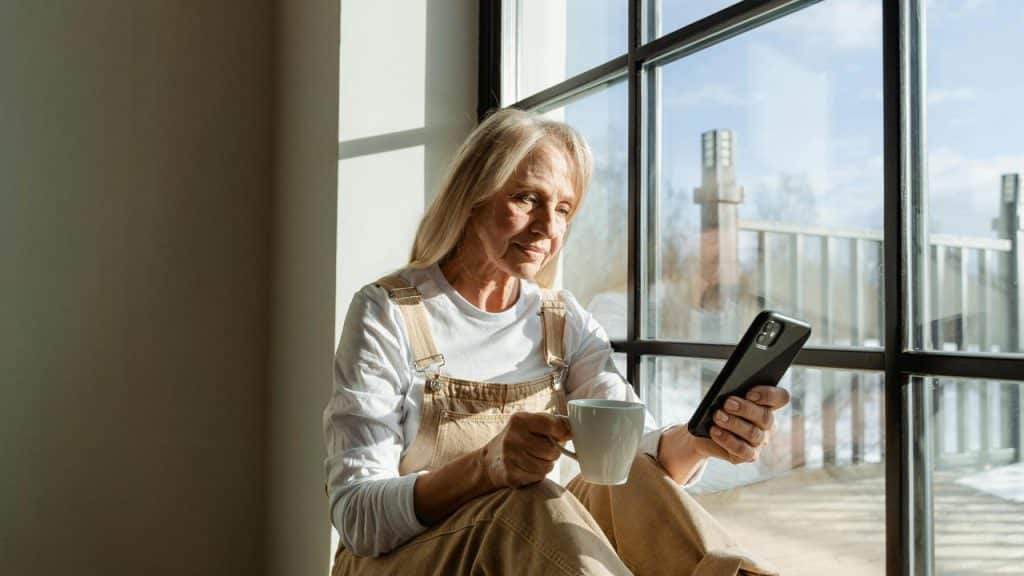 A senior woman sitting by a window holding a cup and looking at her phone.