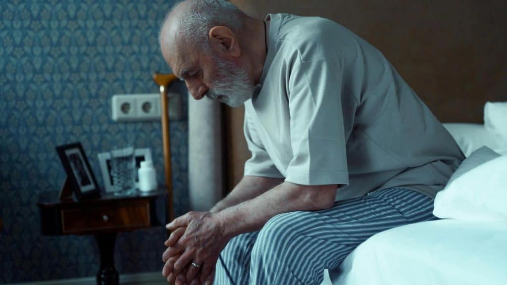 A senior man sitting on the edge of a bed with a thoughtful expression.