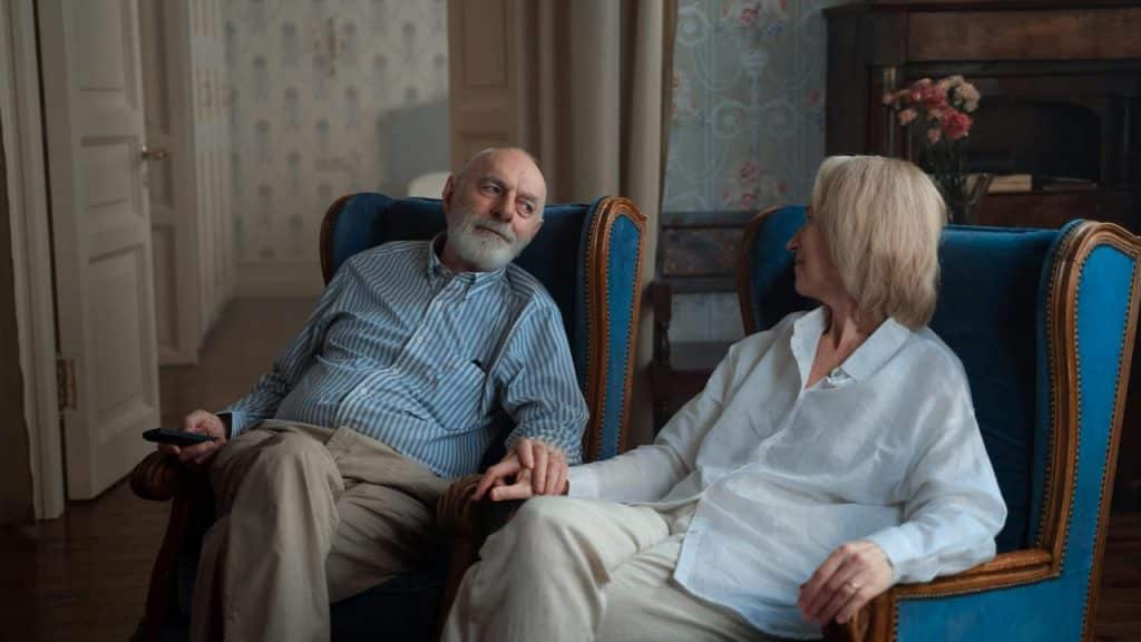 A senior couple sitting on armchairs holding hands and looking at each other.