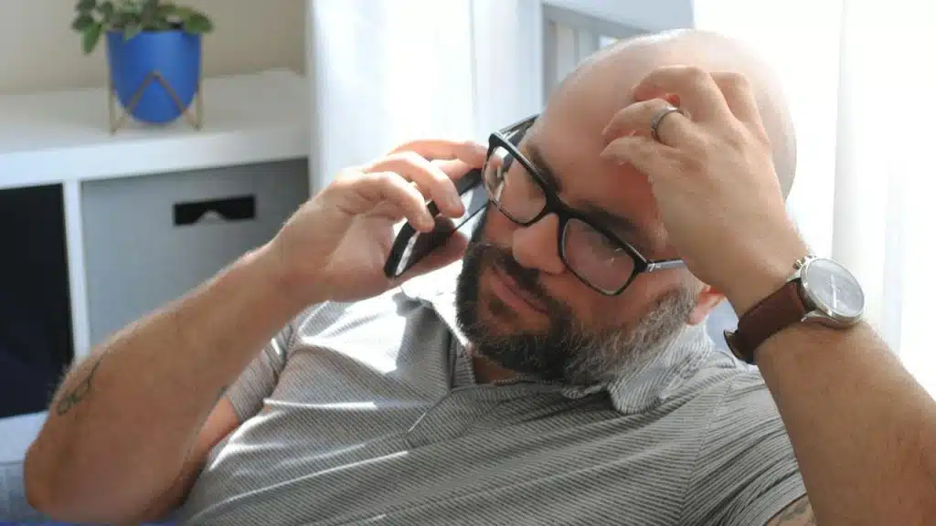 A man wearing glasses talking on the phone while looking stressed and holding his head.