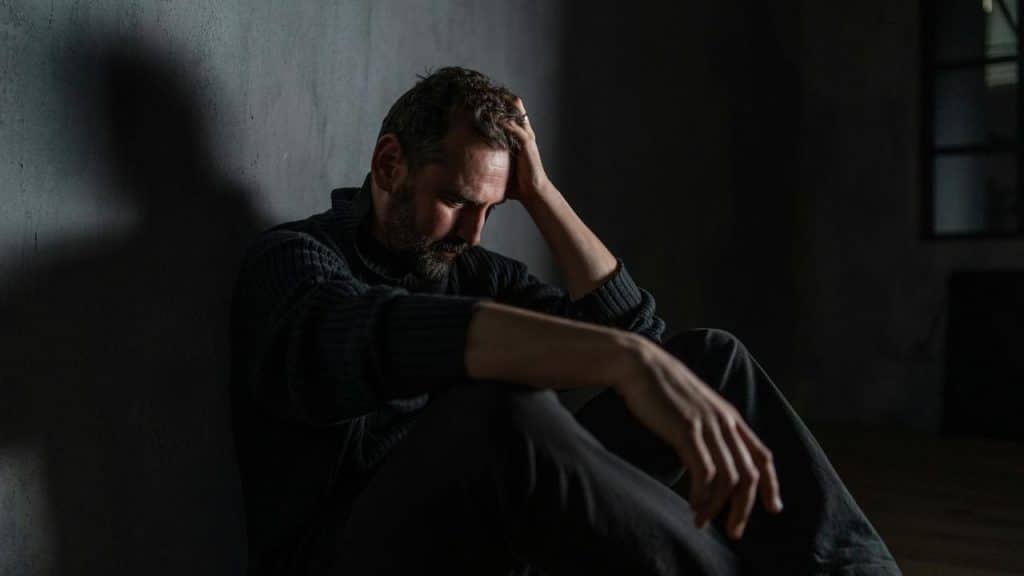 A man sitting against a wall with his head in his hand, looking distressed.