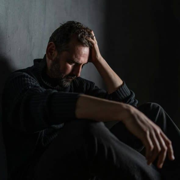 A man sitting against a wall with his head in his hand, looking distressed.