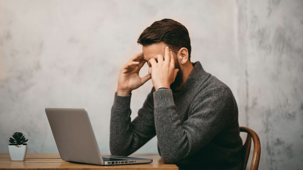 A man sits at a table with a laptop, holding his head in his hands.