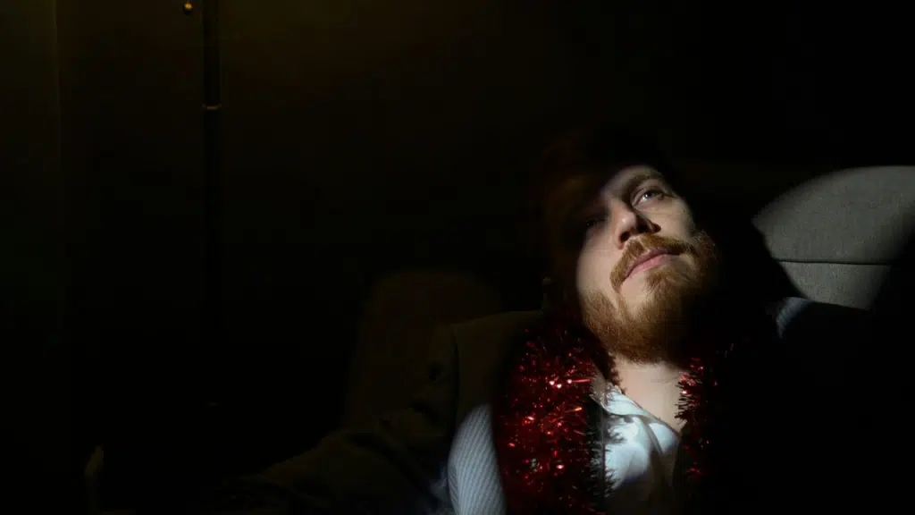 A man with a beard and a red tinsel garland looks up while sitting in a dark room.