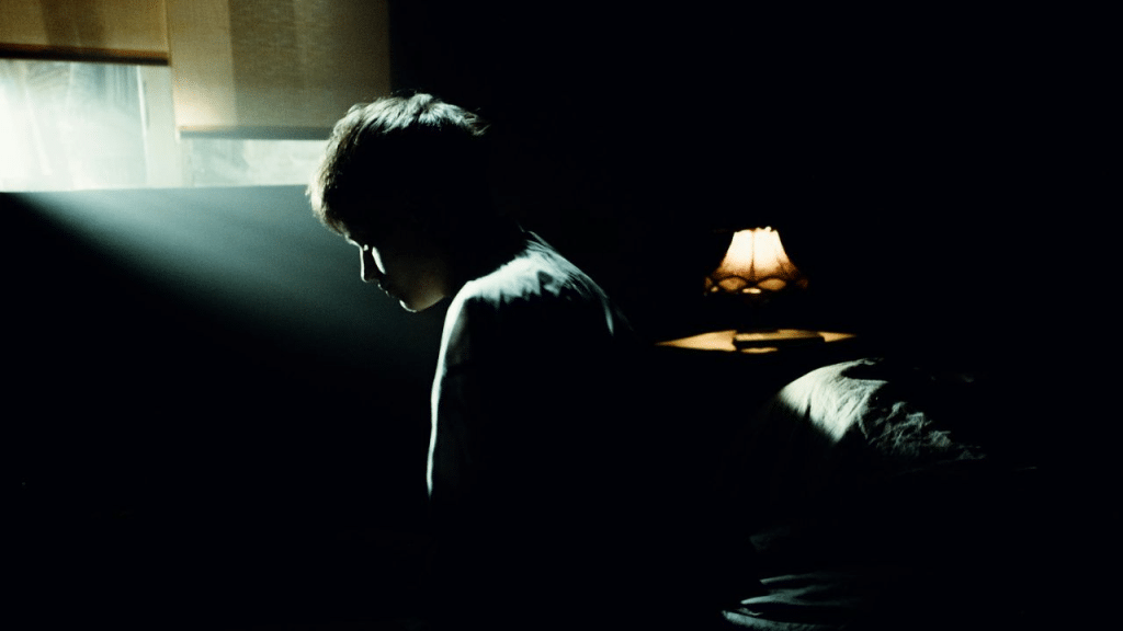 A person sits alone on a bed in a dark room with a lamp on a nightstand.