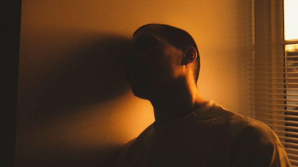 A silhouette of a person stands against a wall next to a window.