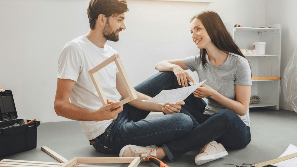 A happy couple sits on the floor assembling furniture, looking at each other and smiling.