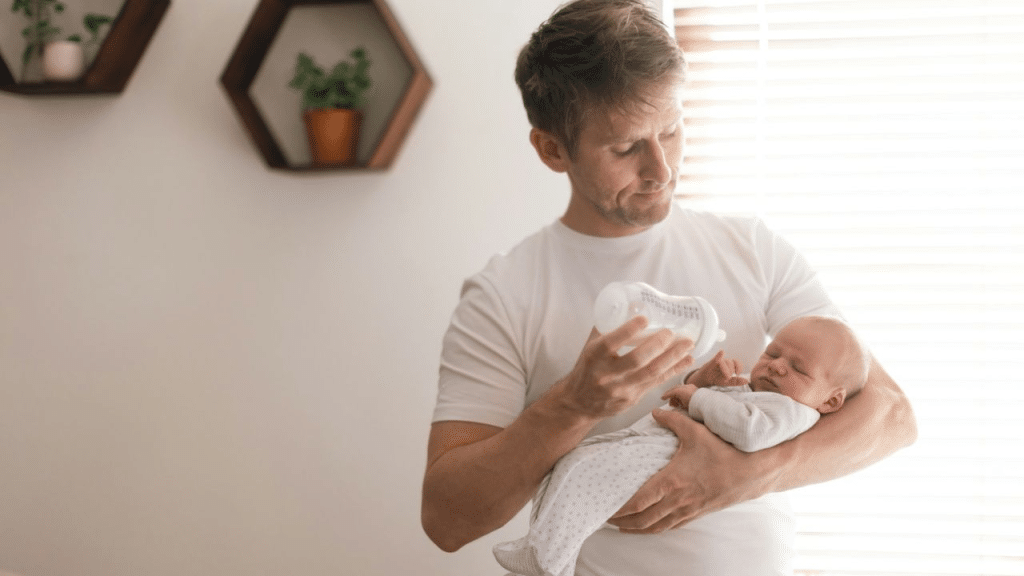 A man in a white T-shirt holds and bottle-feeds a sleeping baby.