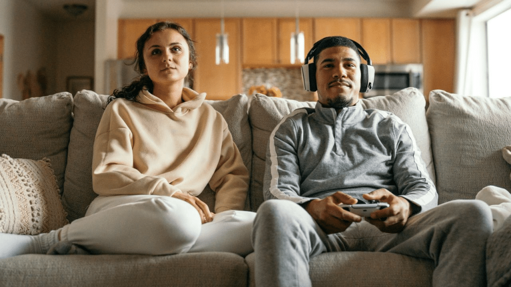 A woman and a man sit on a couch. The man is holding a video game controller.
