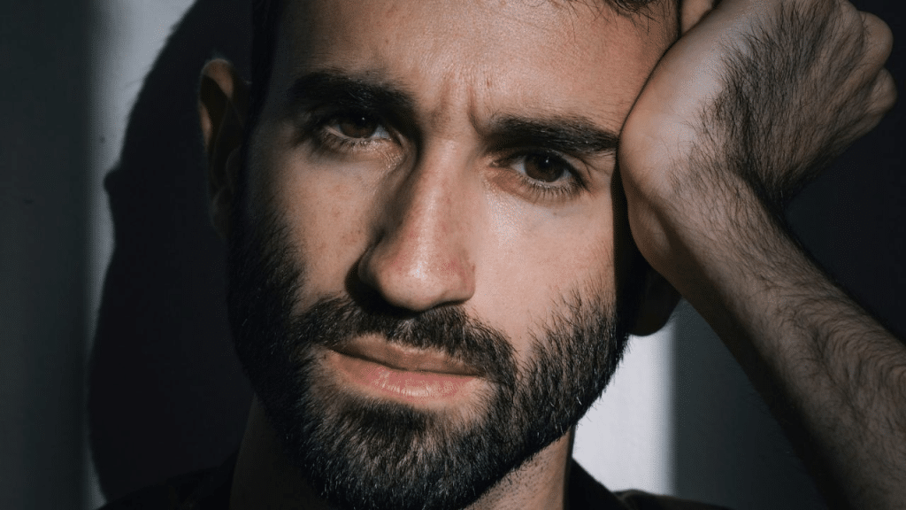 A close-up headshot of a man with a beard, holding his head in his hand.