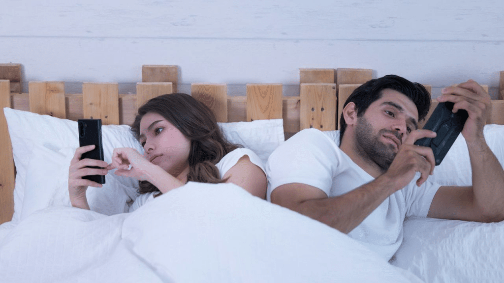 A man and woman lie in bed, back to back, each looking at their phone.