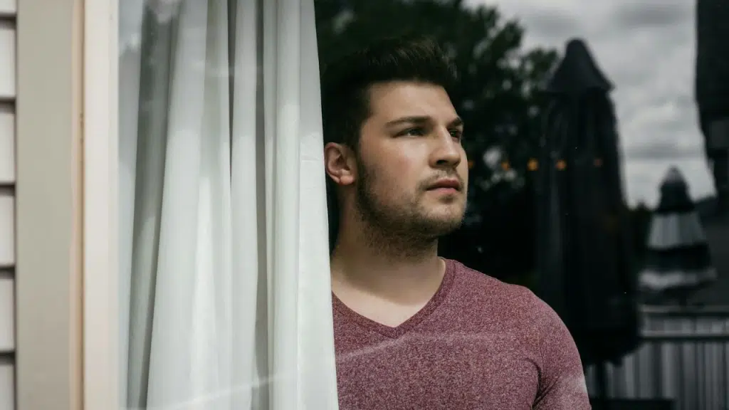 A man looks out a window, half-hidden by a white curtain.