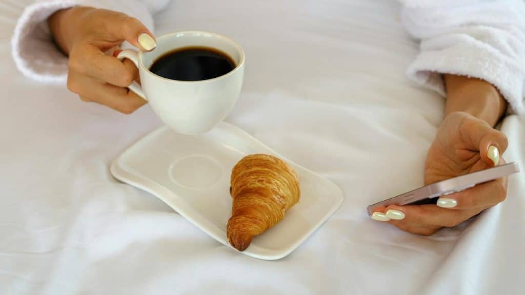A person holding coffee and a phone in bed.