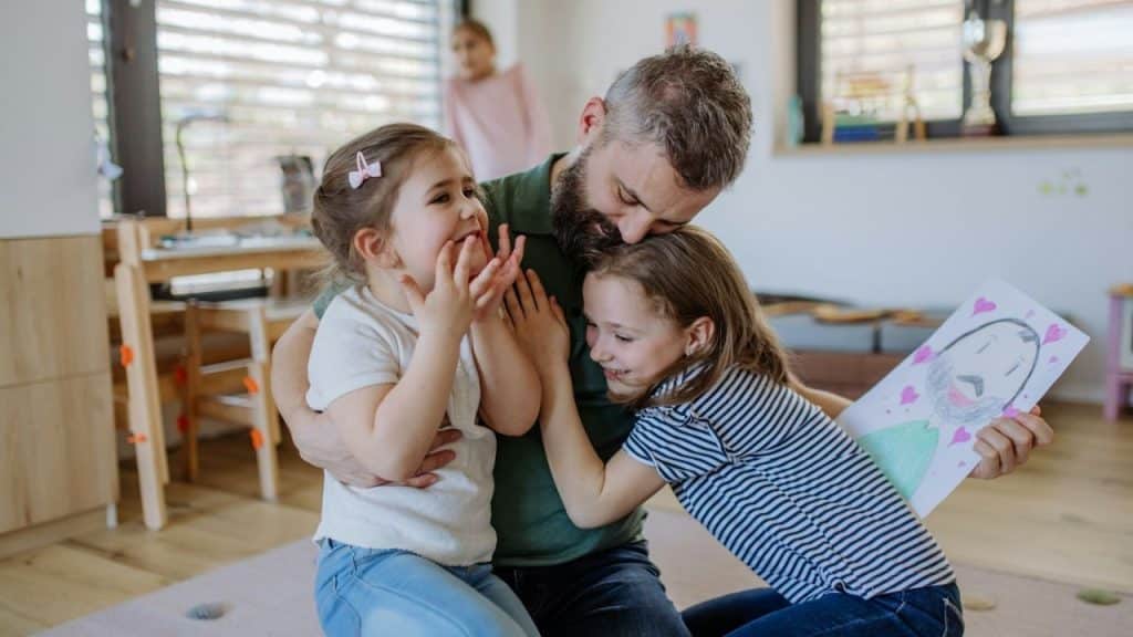 A smiling man hugs two young girls, one of whom holds a drawing of a man with a beard.