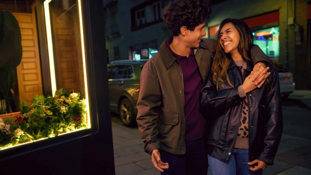 A smiling, young couple walking down a city street at night with the man's arm around the woman's shoulder.