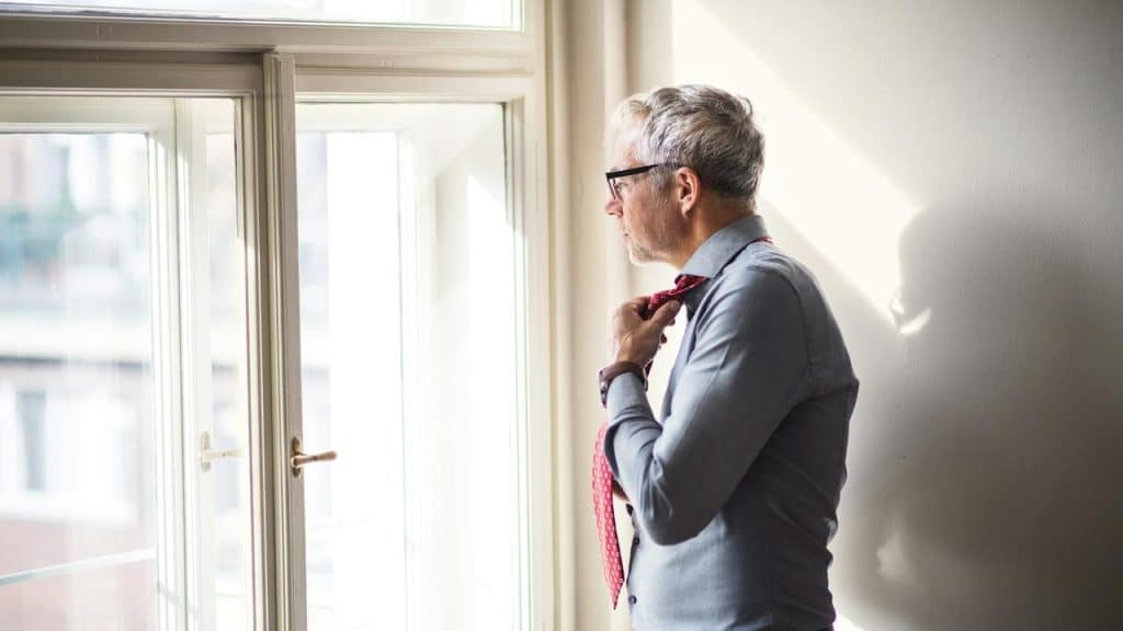 A man standing by a window adjusting his tie.