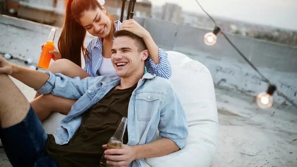 A young couple relaxes on a rooftop, laughing while holding drinks.