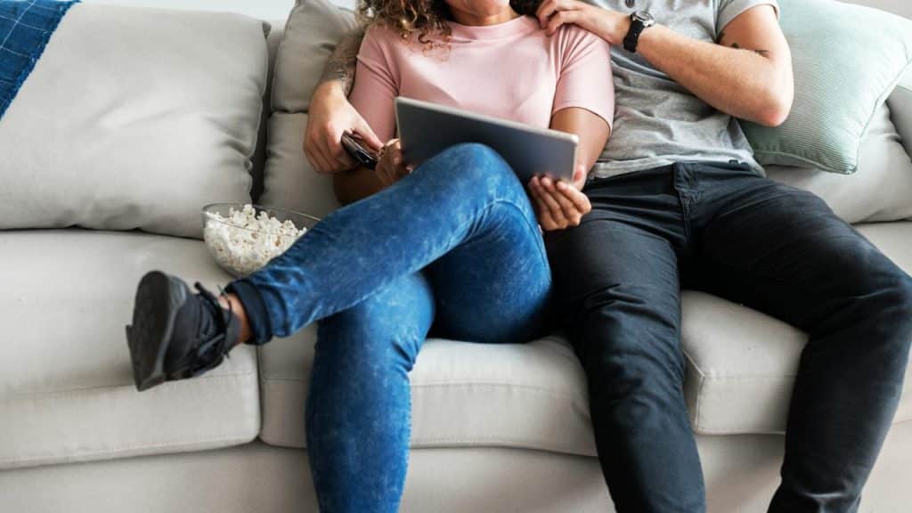 A couple sits on a couch watching something on a tablet with popcorn.