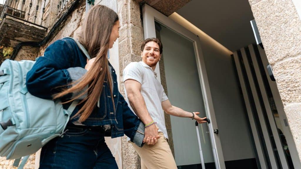 A couple holds hands while arriving at a building with luggage.