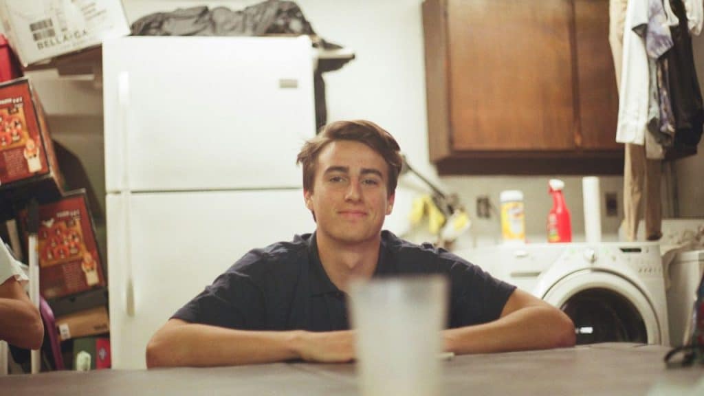 A smiling young man with brown hair sits at a table in a messy kitchen.