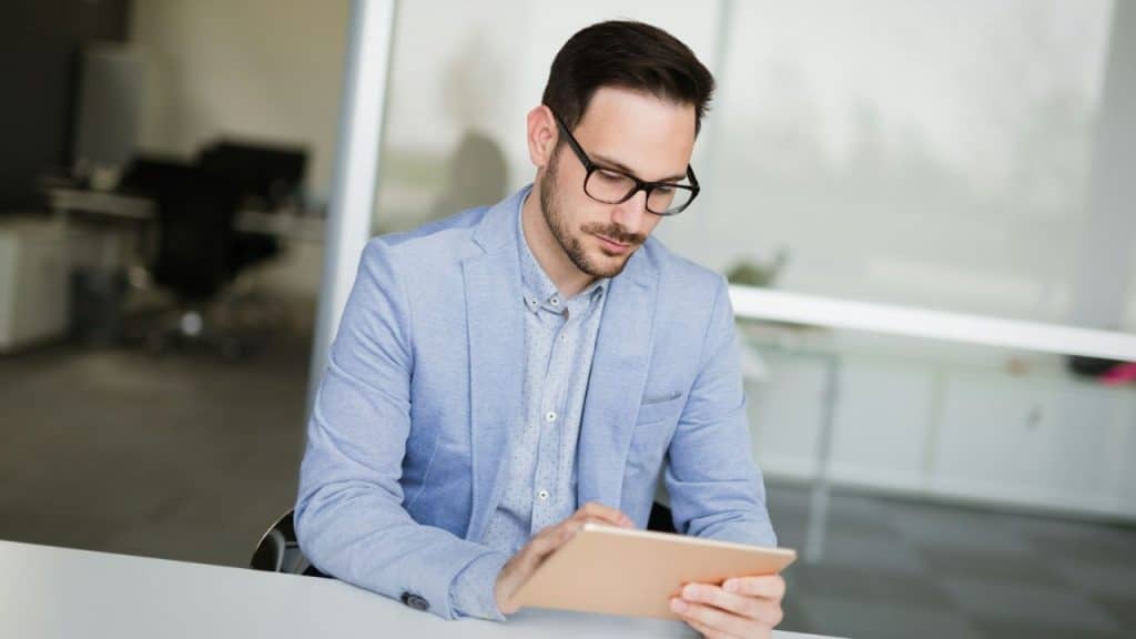 A man wearing glasses and a light blue jacket looks down at a tablet.