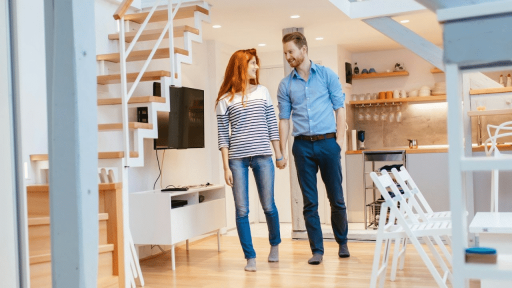 A smiling couple holding hands, walking in their modern, light-filled living room.