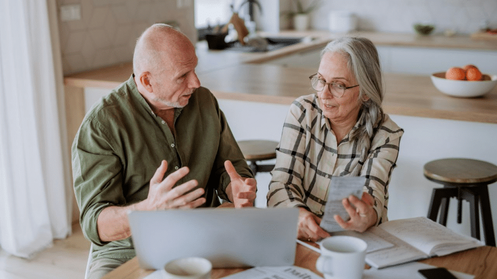 An older man and woman sit at a table, discussing papers and a laptop.