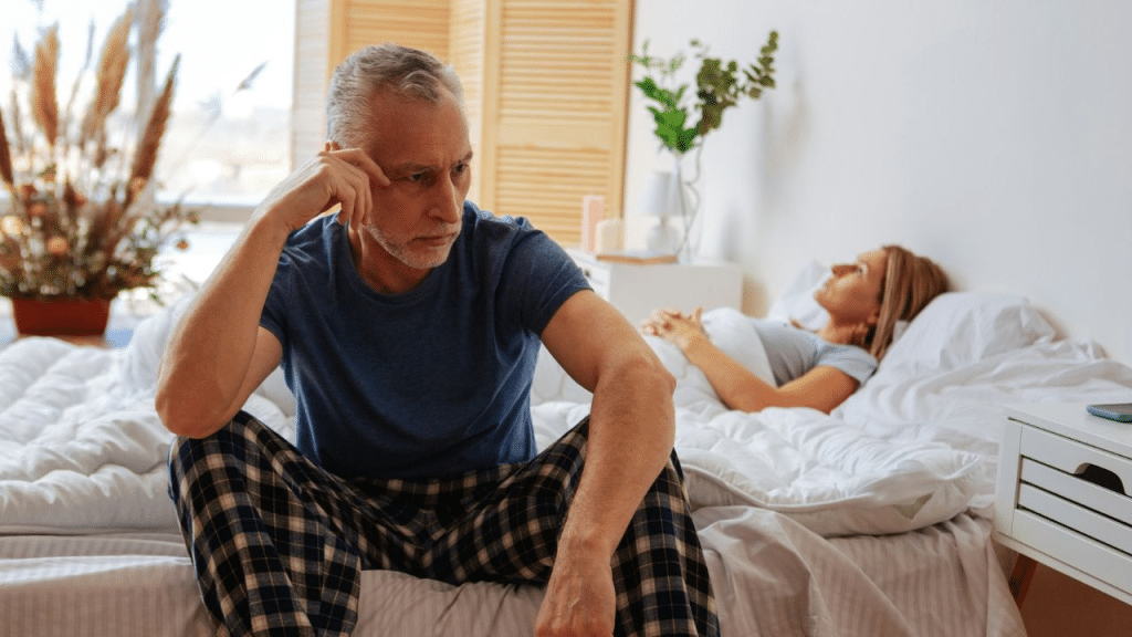 A middle-aged man sits on the bed looking distressed while his wife sleeps.