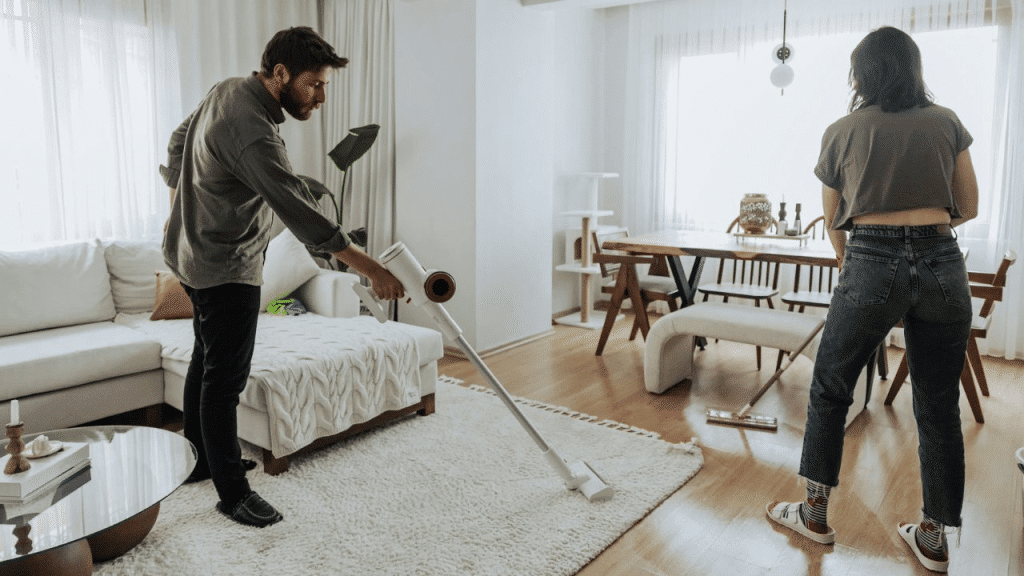 A man and woman sharing household chores, vacuuming and sweeping the floor in a living room.