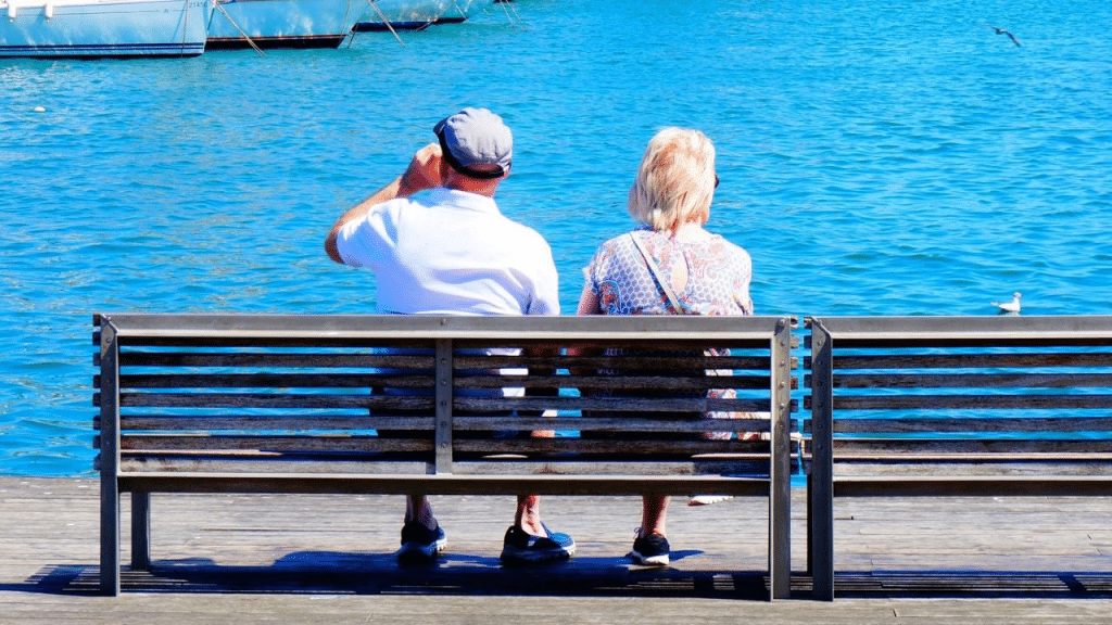 An elderly couple sitting on a bench overlooking the calm blue sea.