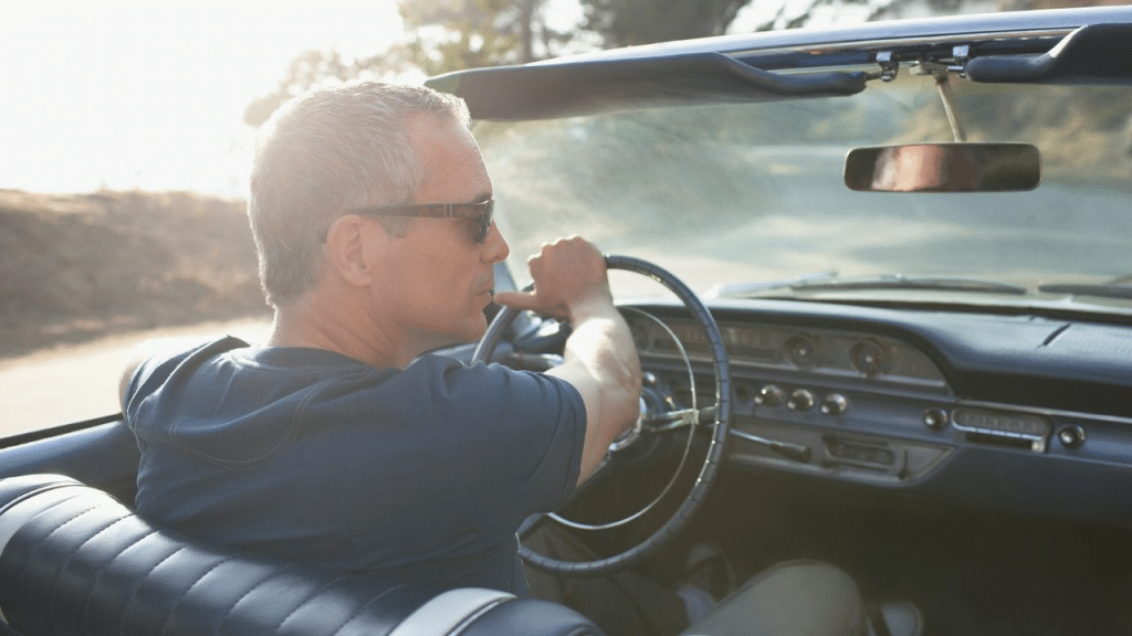 A mature man wearing sunglasses driving a convertible car on a sunny day.