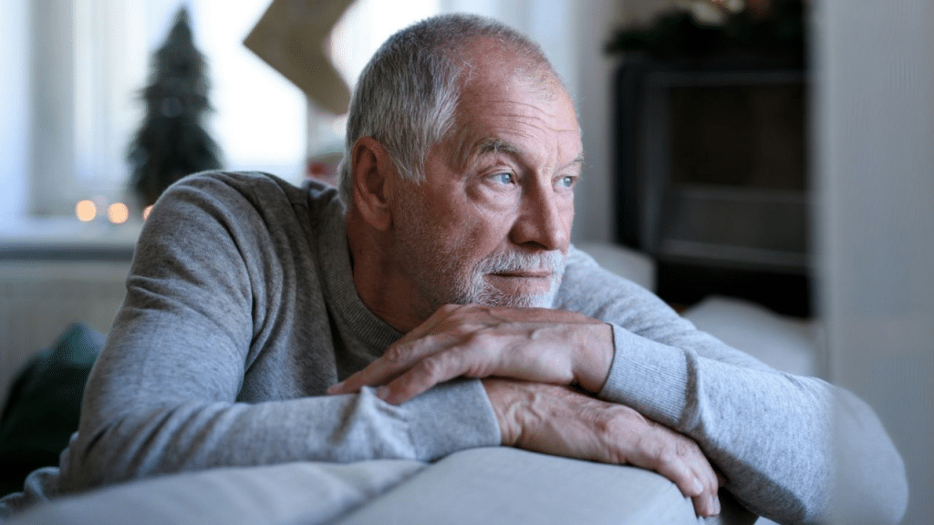 A contemplative older man with a beard leans on a couch, looking away.