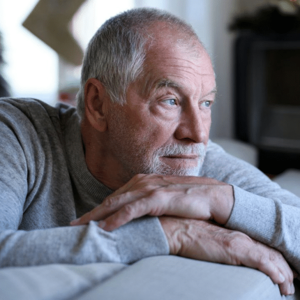 A contemplative older man with a beard leans on a couch, looking away.