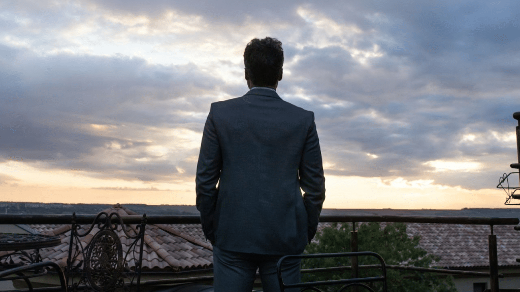 A man in a suit stands on a balcony, looking out at a cloudy sunset.
