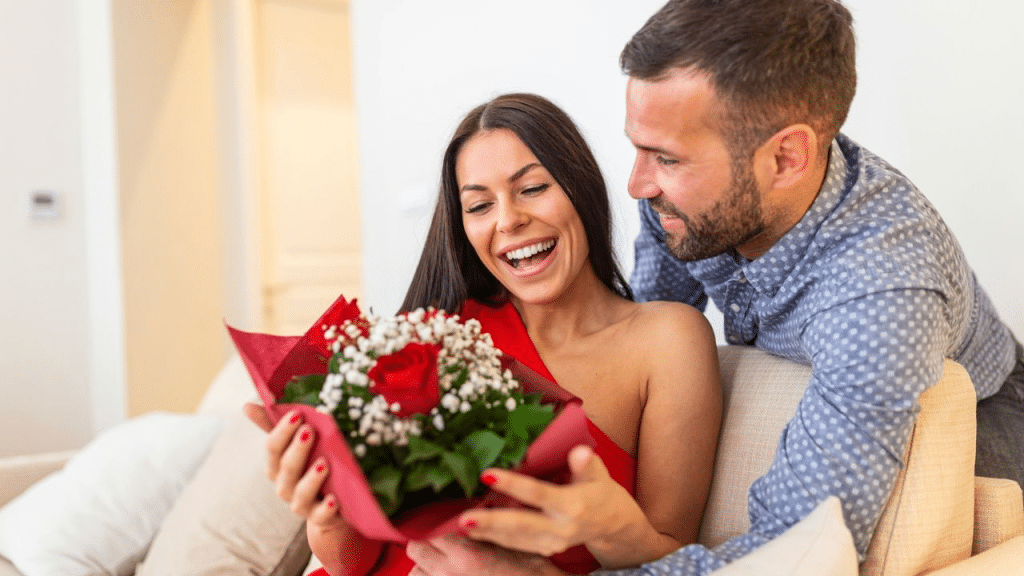 A surprised and happy woman in a red dress receiving a bouquet of flowers from a man.