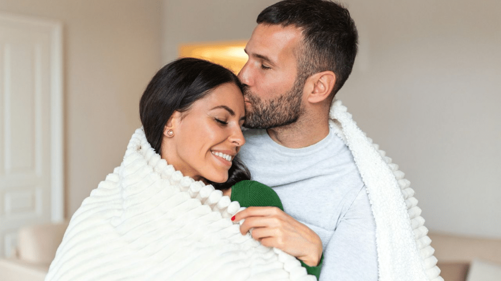 A happy man kissing a smiling woman on her forehead under a cozy blanket.