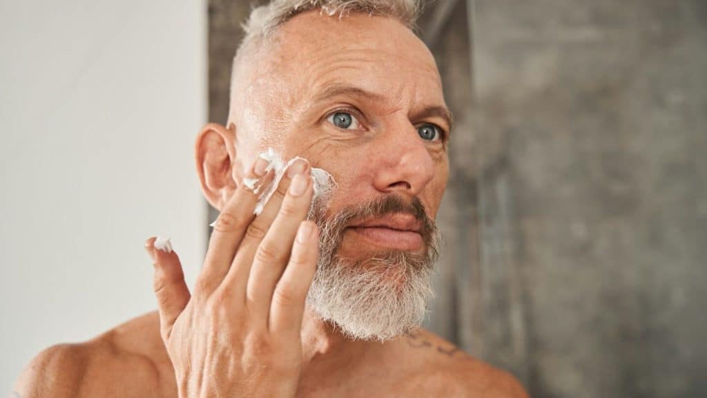 A middle-aged man with a gray beard applies a white cream to his face.